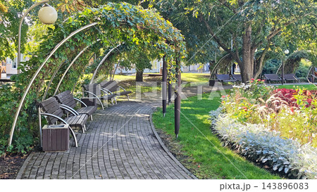 Curving brick walkway with benches beneath leafy trees, arched garden lights, green lawn, and colorful flower beds in a tranquil landscaped public park setting outdoors Curving brick walkway with benches beneath leafy trees, arched garden lights, green lawn, and colorful flower beds in a tranquil landscaped public park setting outdoors 143896083