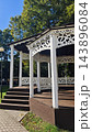 Ornate white wooden gazebo with decorative cutout railings, dark roof, steps, and open sides among mature green trees beside a paved walkway in a landscaped park setting 143896084