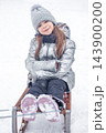 Little girl smiling on a wooden sled in snow wearing a silver winter coat and knit hat 143900200