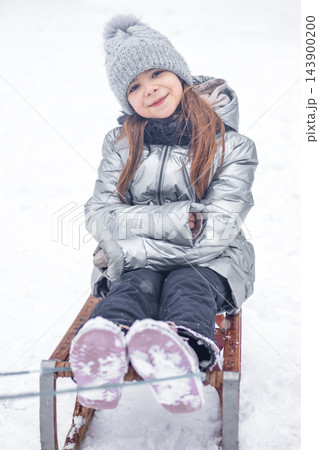 Little girl smiling on a wooden sled in snow wearing a silver winter coat and knit hat Little girl smiling on a wooden sled in snow wearing a silver winter coat and knit hat 143900200