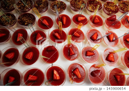 Overhead view of rows of panna cotta cups topped with strawberry sauce and halved strawberries on white surface, some cups finished with crushed nuts and small clear spoons 143900654