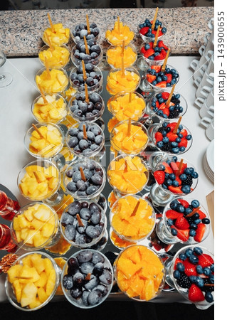 Rows of dessert glasses filled with pineapple grapes oranges and mixed berries on mirrored buffet display with wooden picks for serving at event table arrangement Rows of dessert glasses filled with pineapple grapes oranges and mixed berries on mirrored buffet display with wooden picks for serving at event table arrangement 143900655