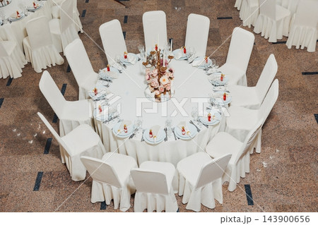Round banquet table with white covered chairs gold candelabra and pink floral centerpiece set for formal dinner event in reception hall with elegant tableware and linens 143900656