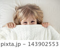 Portrait little girl peeking out blanket on bed. Cute kid smiling hiding under knitted cover. Playful mischievous eye. Hide-and-seek. Children having fun playing active games. Happy childhood concept 143902553