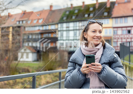 Smiling woman holding smartphone stands on bridge in historic European town, wearing warm jacket and scarf, enjoying calm urban mood. Image represents modern communication, travel lifestyle and link Smiling woman holding smartphone stands on bridge in historic European town, wearing warm jacket and scarf, enjoying calm urban mood. Image represents modern communication, travel lifestyle and link 143902562