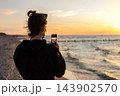 Woman photographing sunset seascape smartphone sandy beach enjoying calm summer evening travel lifestyle leisure moment technology use. Golden hour light sea waves horizon outdoor relaxation scene 143902570