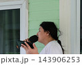 An LGBTQ person dark hair tied back white shirt holds black microphone near their mouth beside green house window white trim framing the outdoor singing activity 143906253