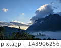 Dark mountain ridges rise above mist filled valley beneath blue sky scattered clouds warm sunlight along the horizon creating broad layered landscape quiet rural detail 143906256