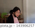 An LGBTQ person dark hair glasses sits indoors wearing pink top dark outer layer lowered face mask while soft light blurred furniture frame an everyday portrait 143906257