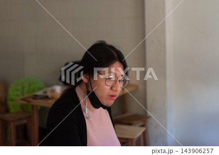 An LGBTQ person dark hair glasses sits indoors wearing pink top dark outer layer lowered face mask while soft light blurred furniture frame an everyday portrait 143906257
