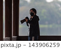 An LGBTQ photographer wearing face mask dark jacket stands beside tall wooden columns holding camera long lens while blurred misty landscape forms the outdoor background 143906259
