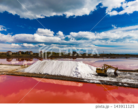 Salt extraction at a pond with an excavator working beside a large salt pile under a cloud-filled sky Salt extraction at a pond with an excavator working beside a large salt pile under a cloud-filled sky 143911125