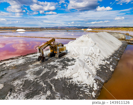 Excavator gathers salt beside pink evaporation ponds at a salt extraction site under a clouded sky Excavator gathers salt beside pink evaporation ponds at a salt extraction site under a clouded sky 143911126