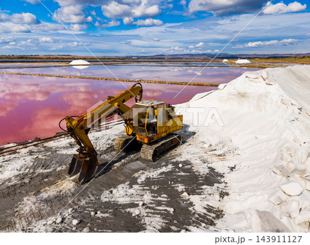 Excavator moves salt beside a pink evaporation pond at a salt extraction site under a cloudy daytime sky Excavator moves salt beside a pink evaporation pond at a salt extraction site under a cloudy daytime sky 143911127