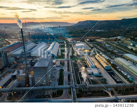 Industrial plant with buildings, storage tanks, pipes, and smoke beside a town at sunset 143911230