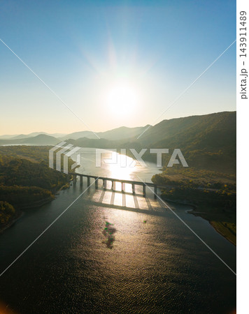 Sunlight reflects across a reservoir crossed by a bridge between wooded hills at sunset 143911489