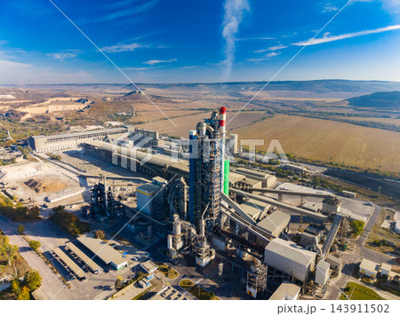 A cement plant with production towers, conveyor systems, and storage buildings in a rural landscape during daylight A cement plant with production towers, conveyor systems, and storage buildings in a rural landscape during daylight 143911502