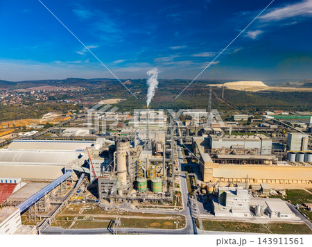 Industrial plant with pipes, buildings, and a plume of steam near hills under a blue sky 143911561