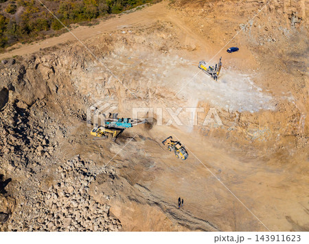 Workers and excavators operate at a quarry as vehicles move across the rock floor during daytime 143911623
