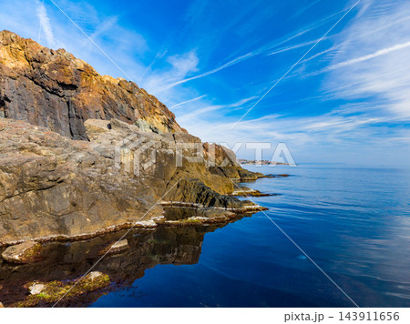 Rocky coast and still sea meet beneath a blue sky during daylight 143911656