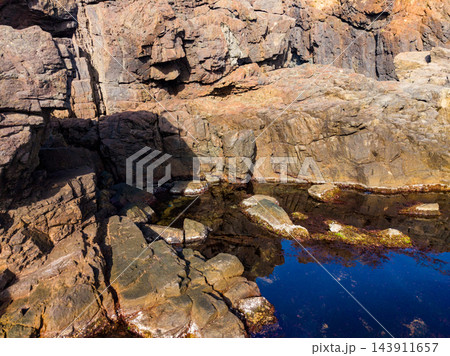 Sunlight falls on a rocky coastal pool beside a layered stone cliff during the day Sunlight falls on a rocky coastal pool beside a layered stone cliff during the day 143911657