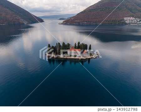 Small island church rests offshore near Perast, Montenegro, surrounded by dark blue water. Aerial landscape includes red roofed stone buildings, cypress trees, distant hills and coastal homes. Small island church rests offshore near Perast, Montenegro, surrounded by dark blue water. Aerial landscape includes red roofed stone buildings, cypress trees, distant hills and coastal homes. 143912798