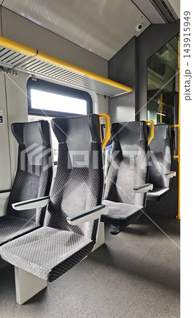 Empty modern public train seats with patterned upholstery, gray flooring, yellow handrails, bright window, and sleek interior fixtures inside a contemporary passenger rail carriage Empty modern public train seats with patterned upholstery, gray flooring, yellow handrails, bright window, and sleek interior fixtures inside a contemporary passenger rail carriage 143915949