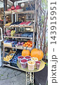 Colorful outdoor fruit and vegetable market stand with pumpkins, apples, oranges, berries, grapes, potatoes, and packaged produce displayed on shelves beside a shop entrance 143915951