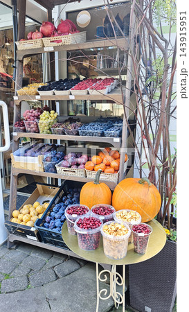 Colorful outdoor fruit and vegetable market stand with pumpkins, apples, oranges, berries, grapes, potatoes, and packaged produce displayed on shelves beside a shop entrance 143915951