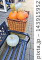 Assorted orange, pale green, and blue pumpkins arranged in a woven basket beside a single white pumpkin on an outdoor market bench near a paved walkway 143915952