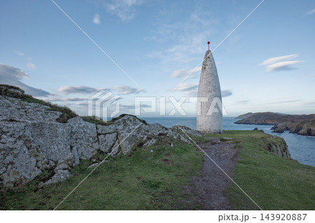 Tall white monument standing on a rocky cliff by the blue sea under a cloudy sky during the daytime. 143920887