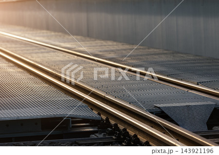Closeup detail railway tracks on industrial steel bridge at sunset, with metal grating and glowing rails fading into distance. Evening sunlight highlights transport infrastructure and geometric lines 143921196