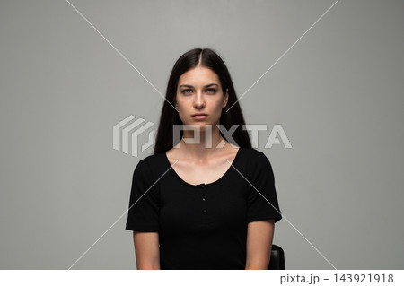 Studio portrait of a young brunette woman with long hair and neutral expression, sitting against a grey background 143921918