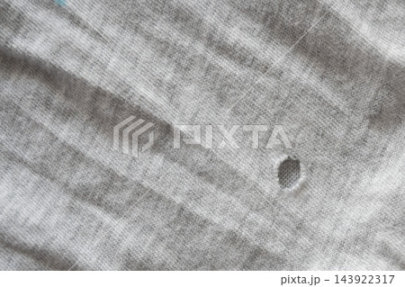 Close-up view of a small hole in a gray cotton t-shirt, highlighting fabric texture, wear, fraying and everyday clothing damage useful for repair, fashion, lifestyle or textile concept images. Close-up view of a small hole in a gray cotton t-shirt, highlighting fabric texture, wear, fraying and everyday clothing damage useful for repair, fashion, lifestyle or textile concept images. 143922317