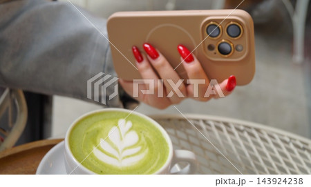 Woman with bright red manicure holds a smartphone above a cup of green matcha latte with leaf foam art. Mobile technology and cafe drink represent connectivity, modern lifestyle and relaxed everyday Woman with bright red manicure holds a smartphone above a cup of green matcha latte with leaf foam art. Mobile technology and cafe drink represent connectivity, modern lifestyle and relaxed everyday 143924238