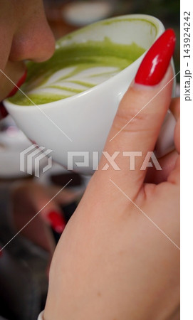 Woman with bright red manicure holds a white cup of green matcha latte with foam art. Warm drink in hand represents comfort, mindful relaxation and contemporary cafe lifestyle 143924242