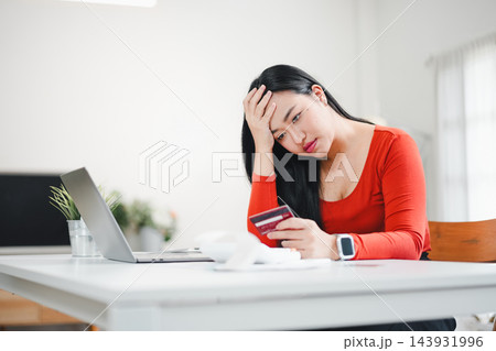 Woman in red shirt worried while checking credit card bill at table with laptop and receipt Woman in red shirt worried while checking credit card bill at table with laptop and receipt 143931996