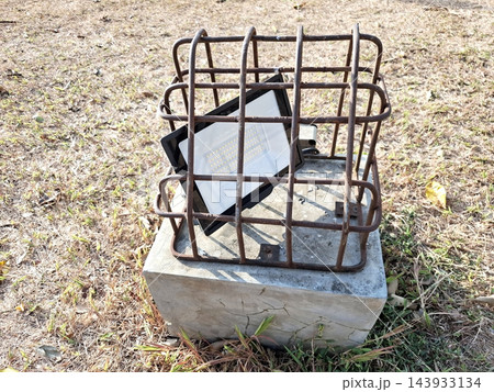 Ground spotlight fixture inside metal cage cage mounted on concrete block. 143933134