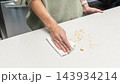 Woman cleaning crumbs from kitchen table after slicing cake, homemade baking process, food preparation routine, cozy home cooking lifestyle. 143934214