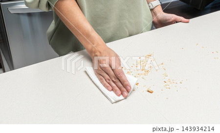 Woman cleaning crumbs from kitchen table after slicing cake, homemade baking process, food preparation routine, cozy home cooking lifestyle. Woman cleaning crumbs from kitchen table after slicing cake, homemade baking process, food preparation routine, cozy home cooking lifestyle. 143934214