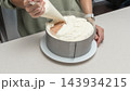 Woman spreading white cream over sponge cake in metal ring on kitchen table, homemade baking process, dessert decoration stage, cozy home cooking lifestyle. 143934215