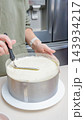 Person in green sweater smooths frosting on a round cake using a spatula in a modern kitchen with stainless steel appliances visible in the background 143934217