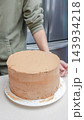 Person decorating a round chocolate cake with smooth frosting on a white cake stand in a modern kitchen setting 143934218