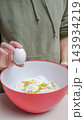 Person holding a white egg above a bowl filled with flour and yolk, wearing a green hoodie in a kitchen setting 143934219