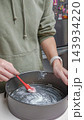 Person in green hoodie applies butter to a round baking pan with a red spatula in a modern kitchen setting with stainless steel appliances 143934220