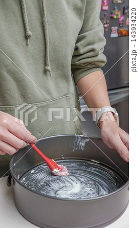 Person in green hoodie applies butter to a round baking pan with a red spatula in a modern kitchen setting with stainless steel appliances Person in green hoodie applies butter to a round baking pan with a red spatula in a modern kitchen setting with stainless steel appliances 143934220