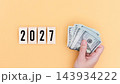 Hand holding a stack of United States dollar bills beside wooden number blocks displaying 2027 year on a warm orange background, financial concept 143934222