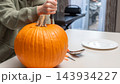 Person carving a large orange pumpkin with a knife on a kitchen table, plate and utensils visible in a well-lit indoor setting 143934227