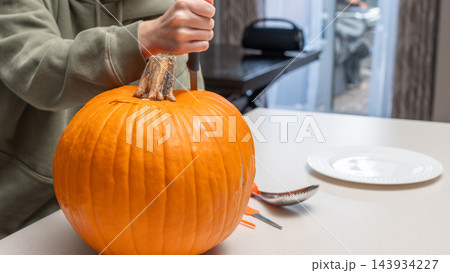 Person carving a large orange pumpkin with a knife on a kitchen table, plate and utensils visible in a well-lit indoor setting Person carving a large orange pumpkin with a knife on a kitchen table, plate and utensils visible in a well-lit indoor setting 143934227