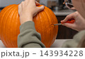 Person carving a bright orange pumpkin for Halloween with a small knife, seated at a table in a cozy indoor setting with kitchen elements visible 143934228